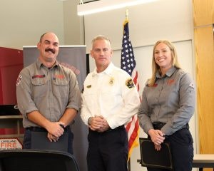 Three people in uniform posing indoors, with an American flag in the background.