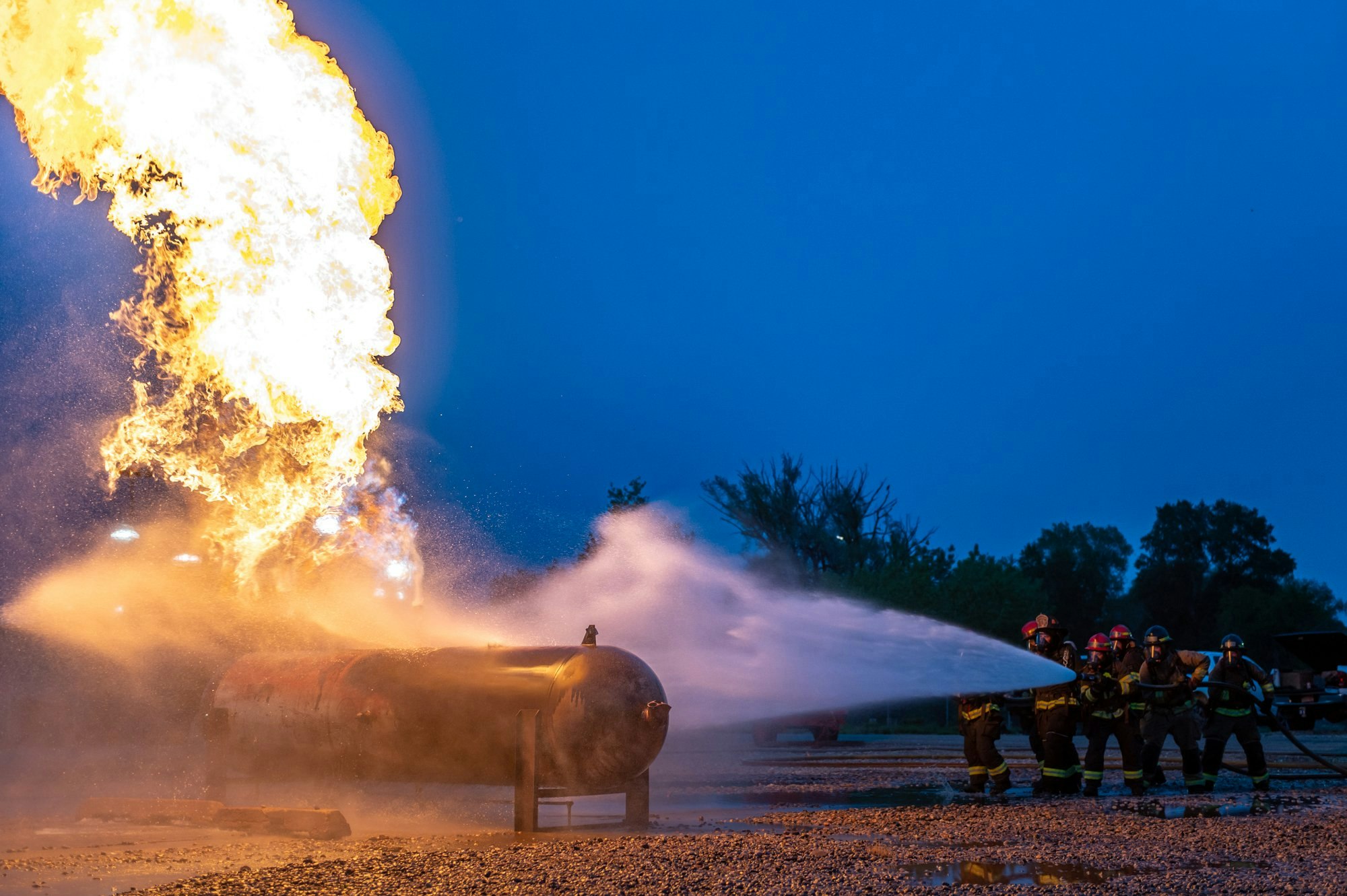 Firefighters battling a large blaze with hoses, spraying water at a burning gas tank.