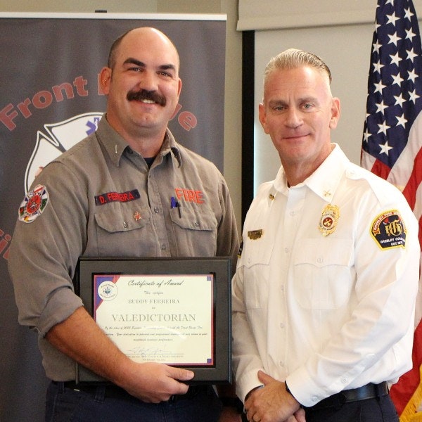 Two men in uniform pose with a "Valedictorian" certificate of award. An American flag is in the background.
