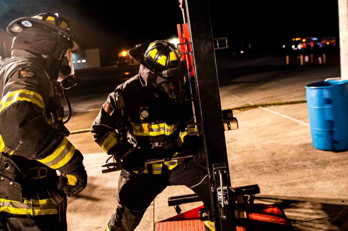 Two firefighters in gear using a tool on a door prop at night.
