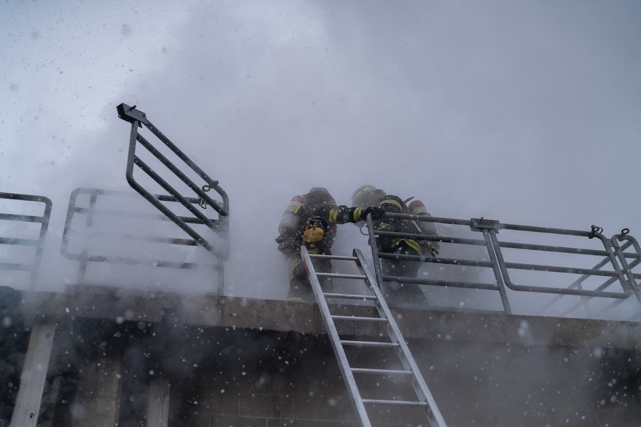 Two firefighters in gear on a smoky rooftop with a ladder and railing, working amidst smoke or mist.