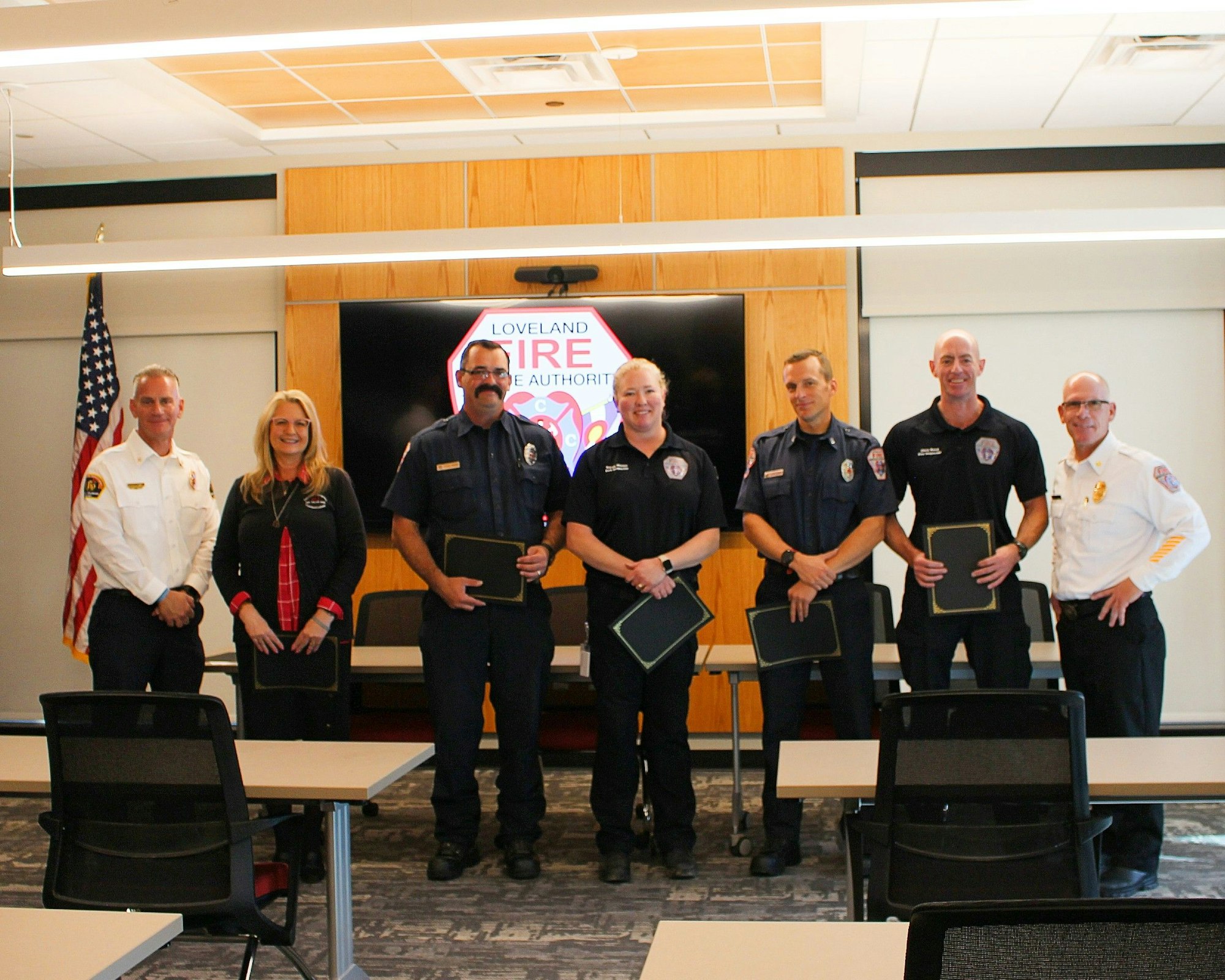 Seven people in uniform holding certificates, standing in a room with a "Loveland Fire" sign and an American flag.