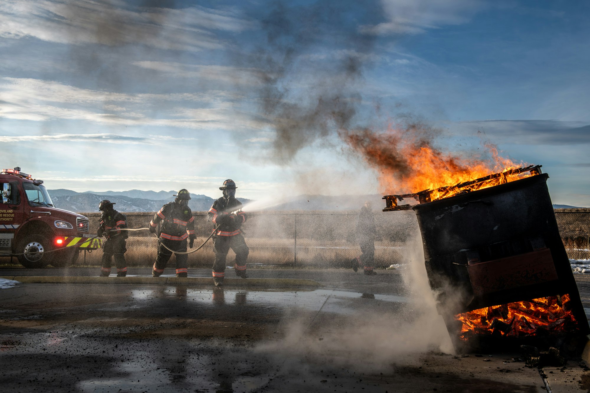 Firefighters extinguishing a large fire with a hose, next to a fire truck, under a cloudy sky.