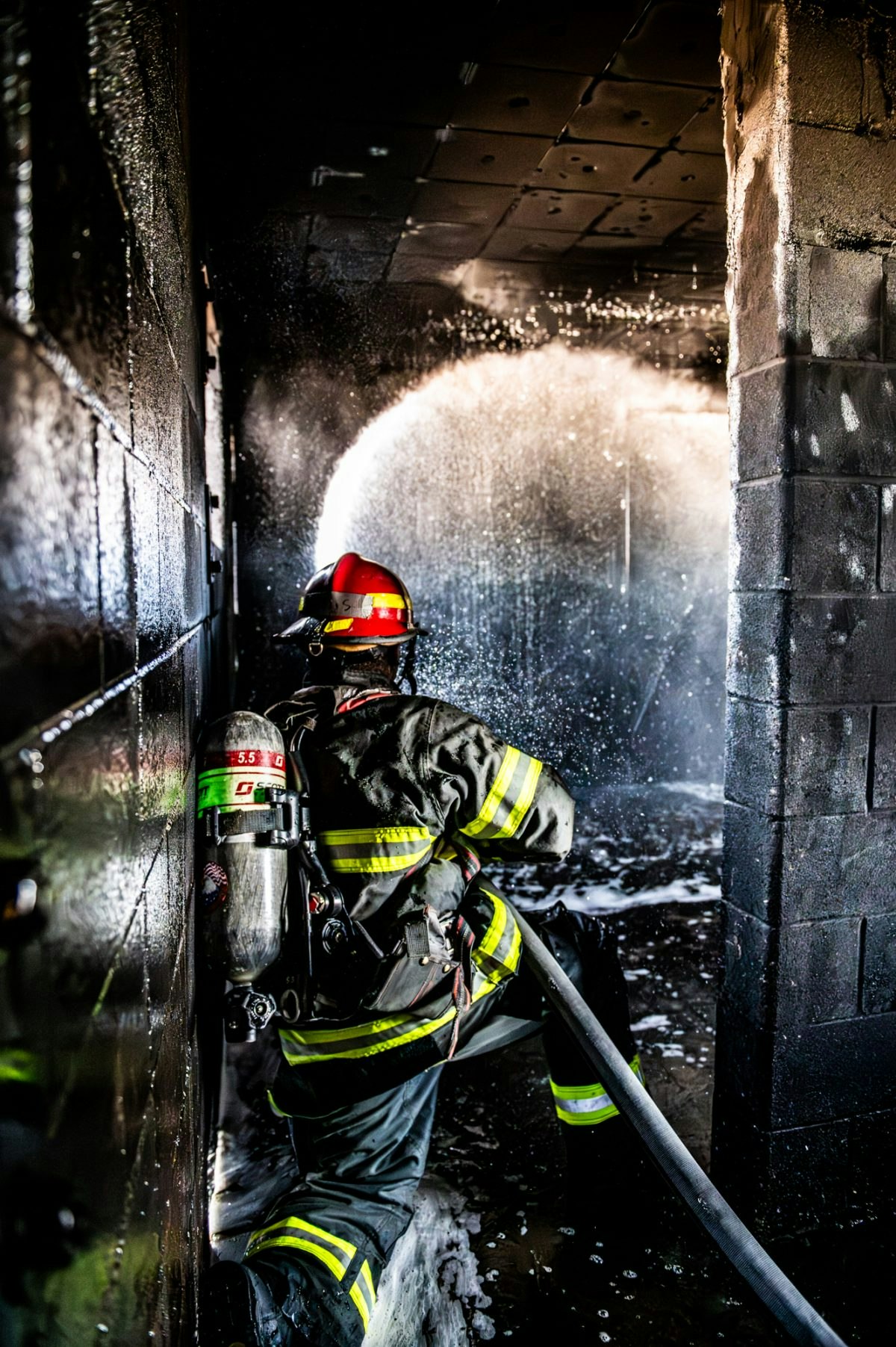 A firefighter in protective gear sprays water from a hose in a dark, smoke-filled environment.