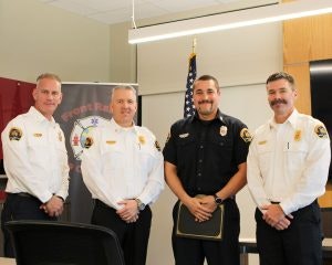 Four men in uniform, three in white and one in black, standing indoors near a flag and fire department banner.