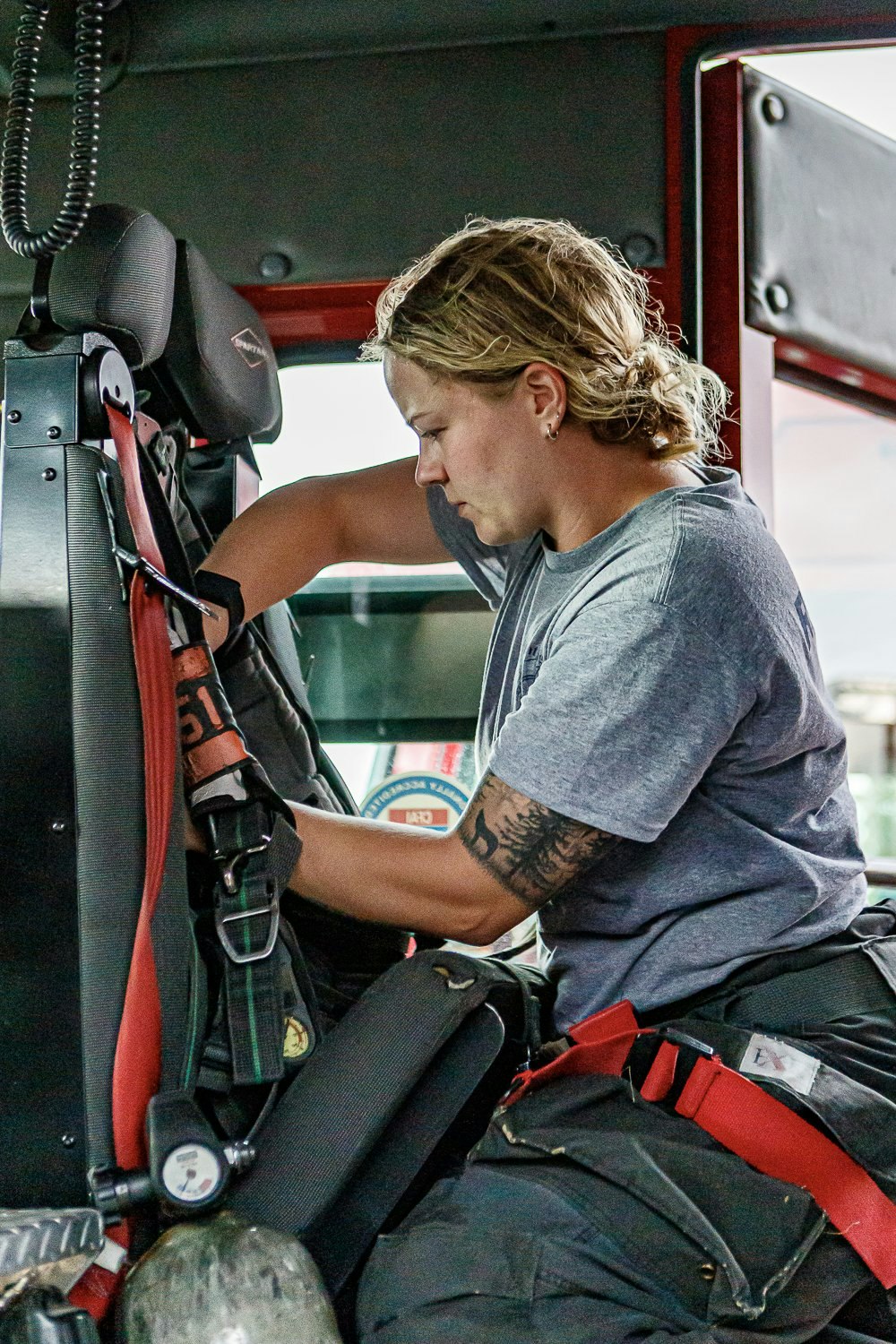 Person sitting in a vehicle, adjusting or securing equipment, wearing a t-shirt and pants with red straps.