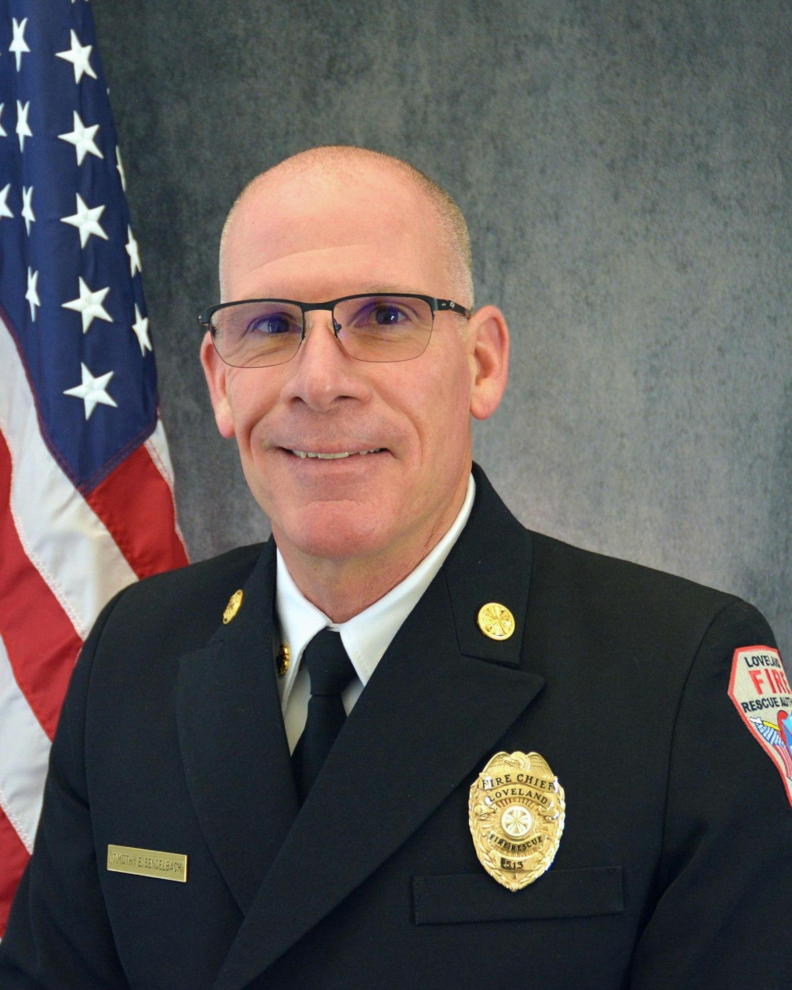 A person in a formal uniform with badges, in front of a U.S. flag, likely a fire department official.