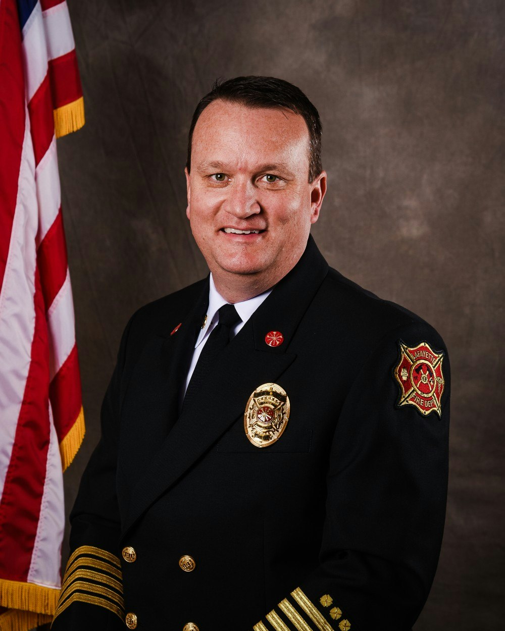 A man in a formal uniform with badges and an American flag backdrop.