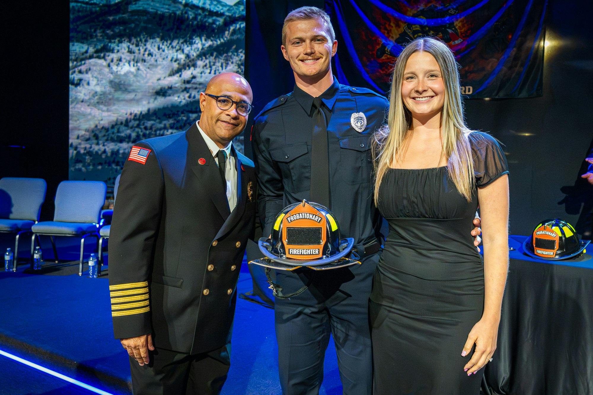 A formal event featuring a new firefighter holding a helmet, flanked by a fire chief and a woman, celebrating his accomplishment.