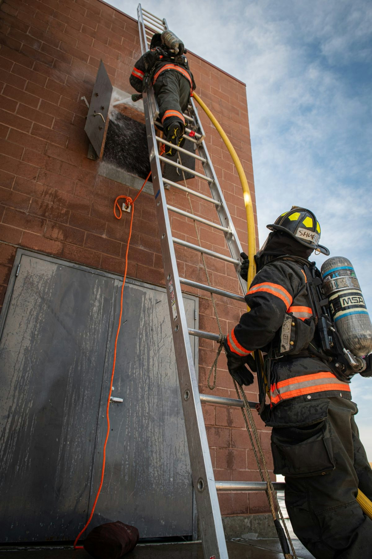 Two firefighters in gear climb a ladder to a window, preparing to extinguish a fire.