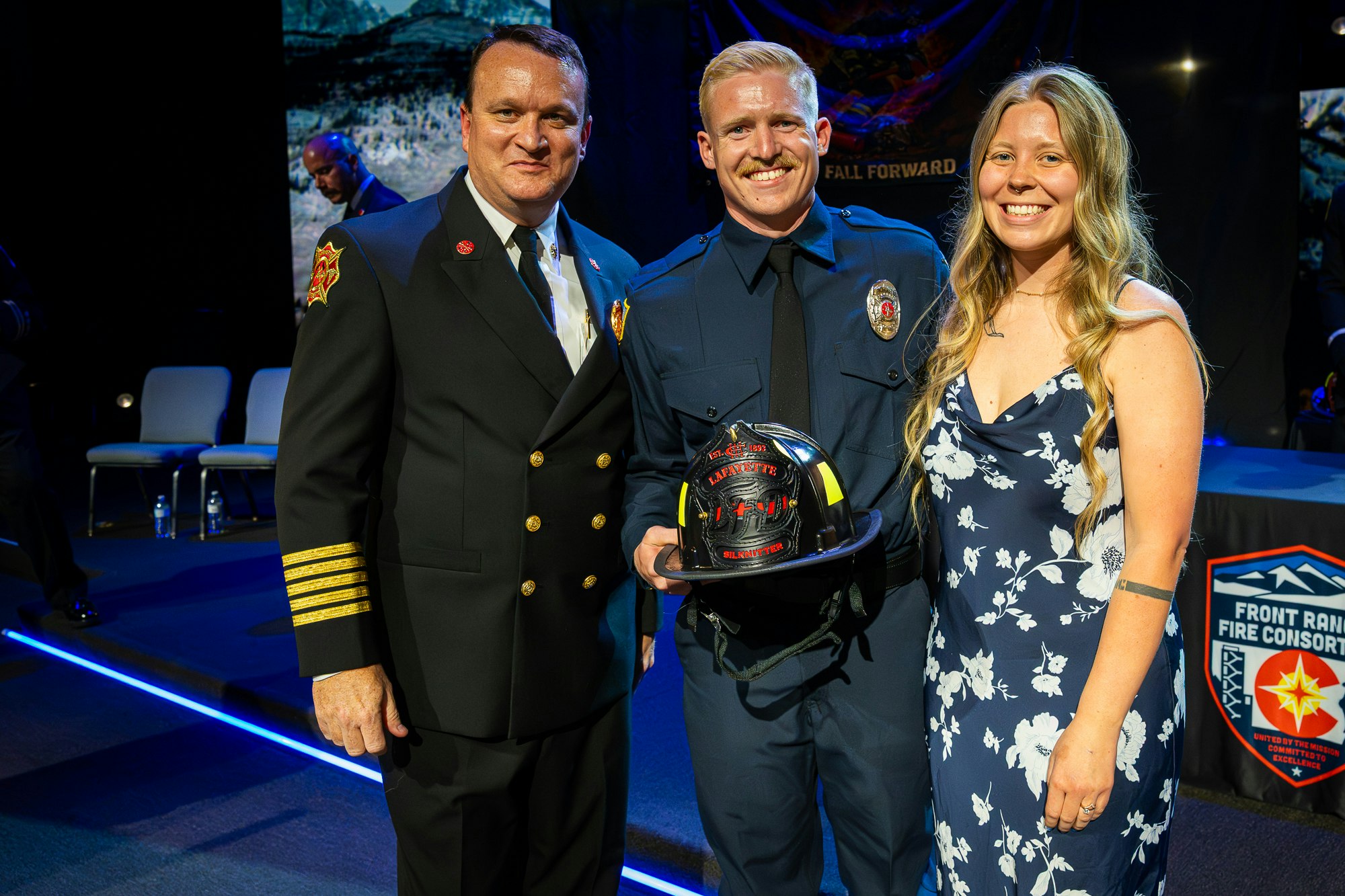 A firefighter smiles with two people during a ceremony, holding a helmet, celebrating a special achievement.