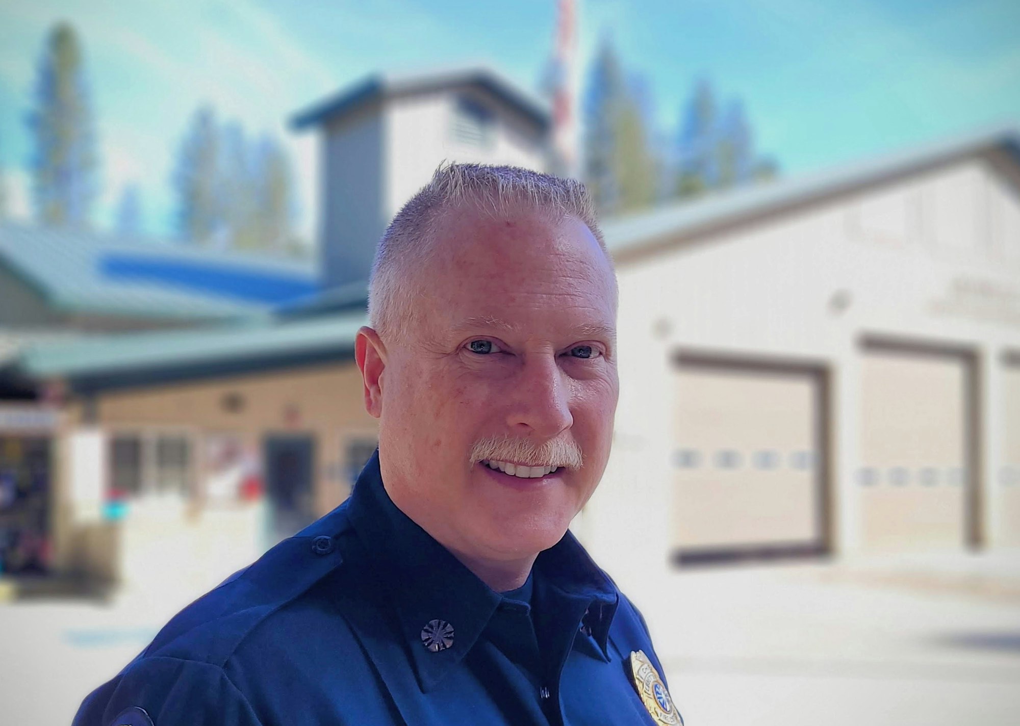 A smiling person in a blue uniform with a blurred background of a building and trees.