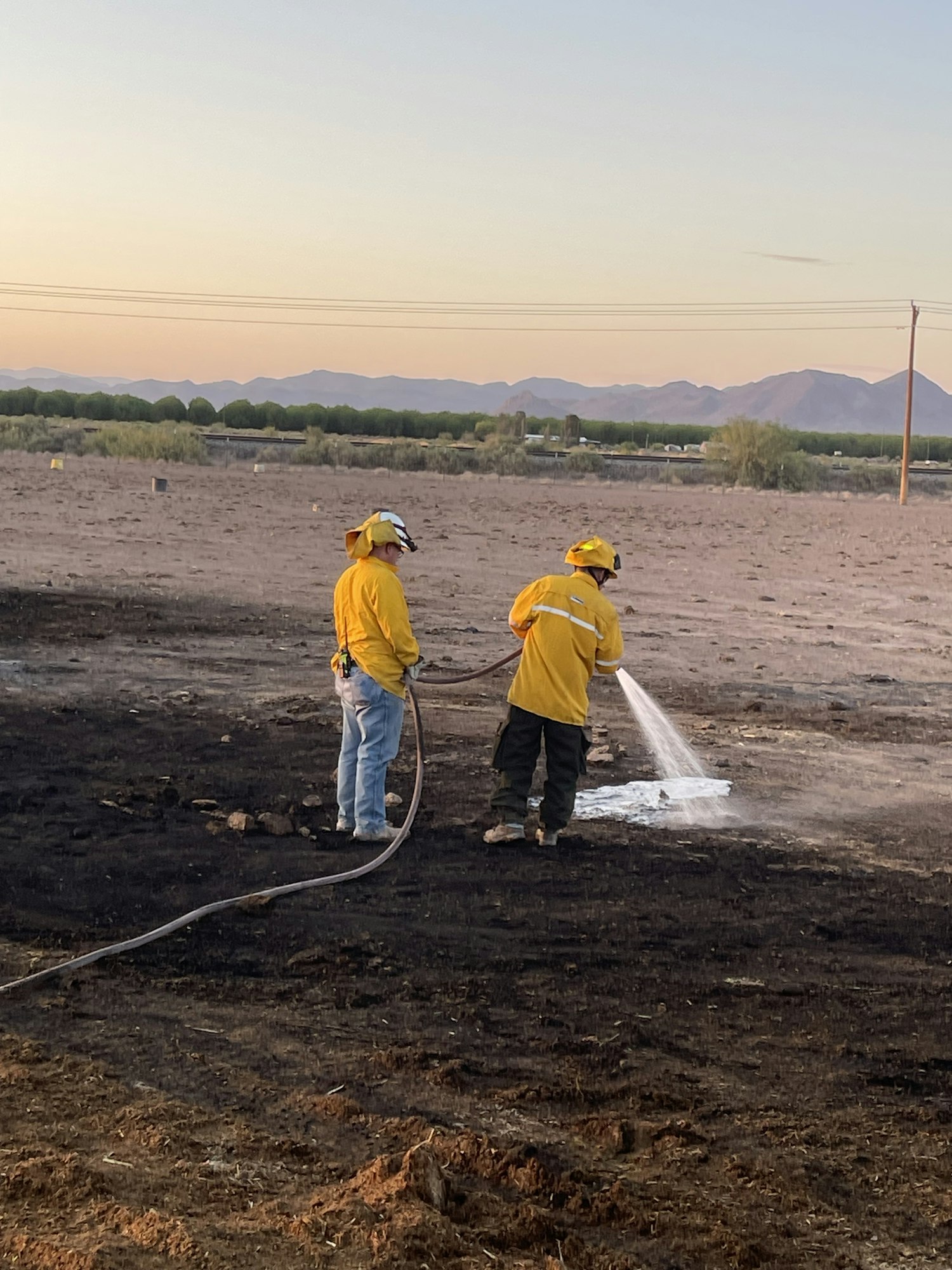 Two people in yellow outfits are spraying water on charred ground, possibly extinguishing a fire.