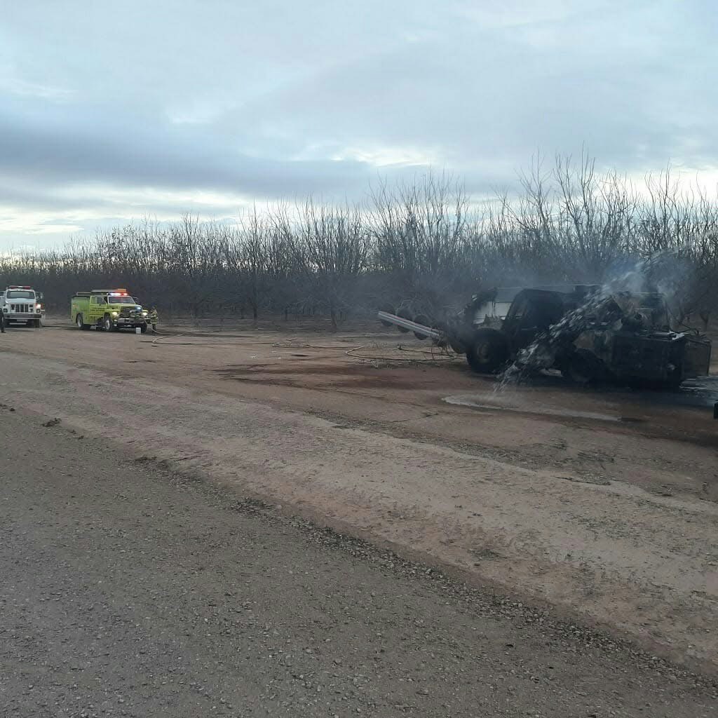 Fire trucks responding to a smoldering vehicle fire on a dirt road with barren trees in the background.