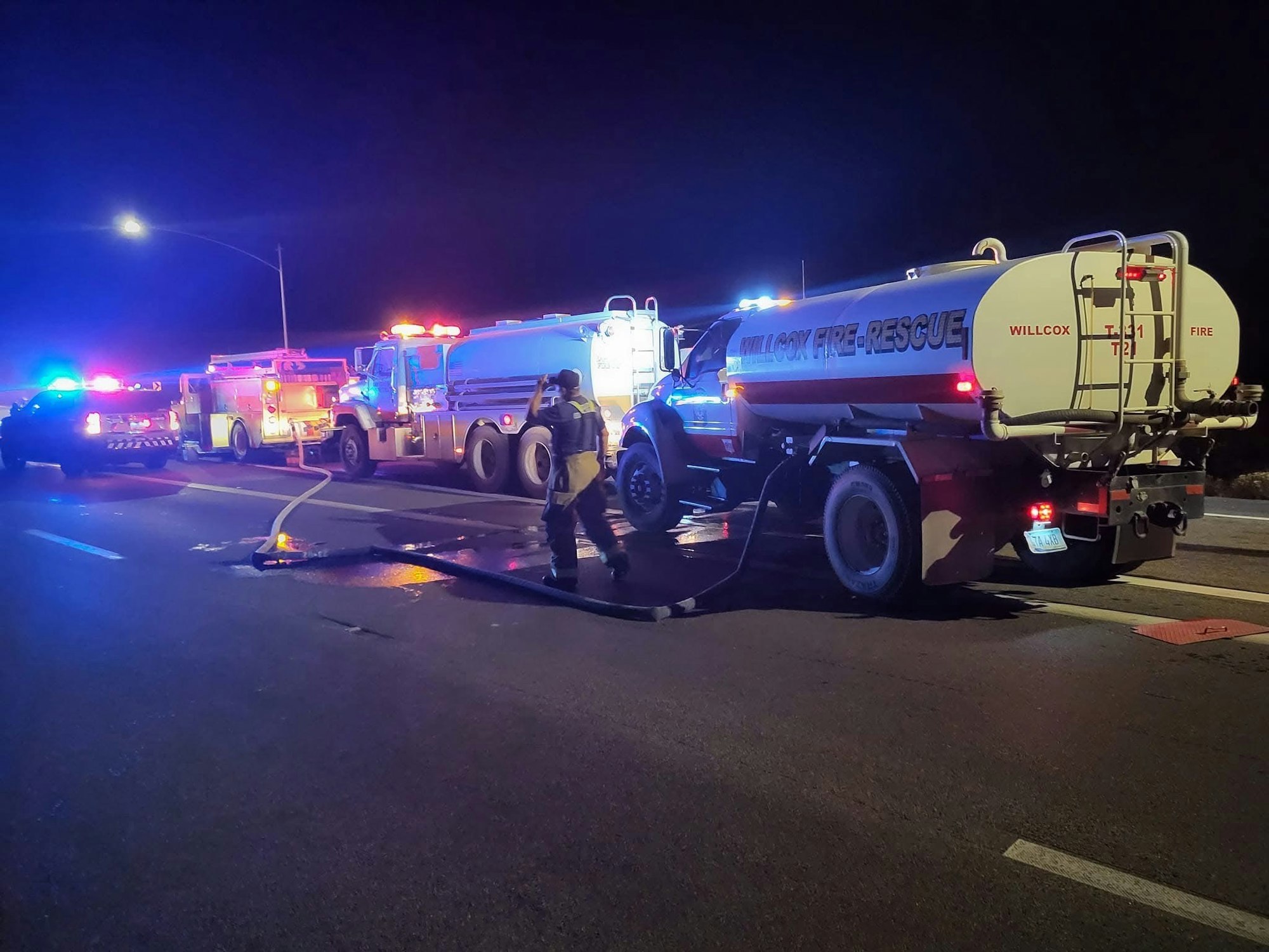 Emergency vehicles on a dark road, with firefighters managing a fire hose, lights flashing.
