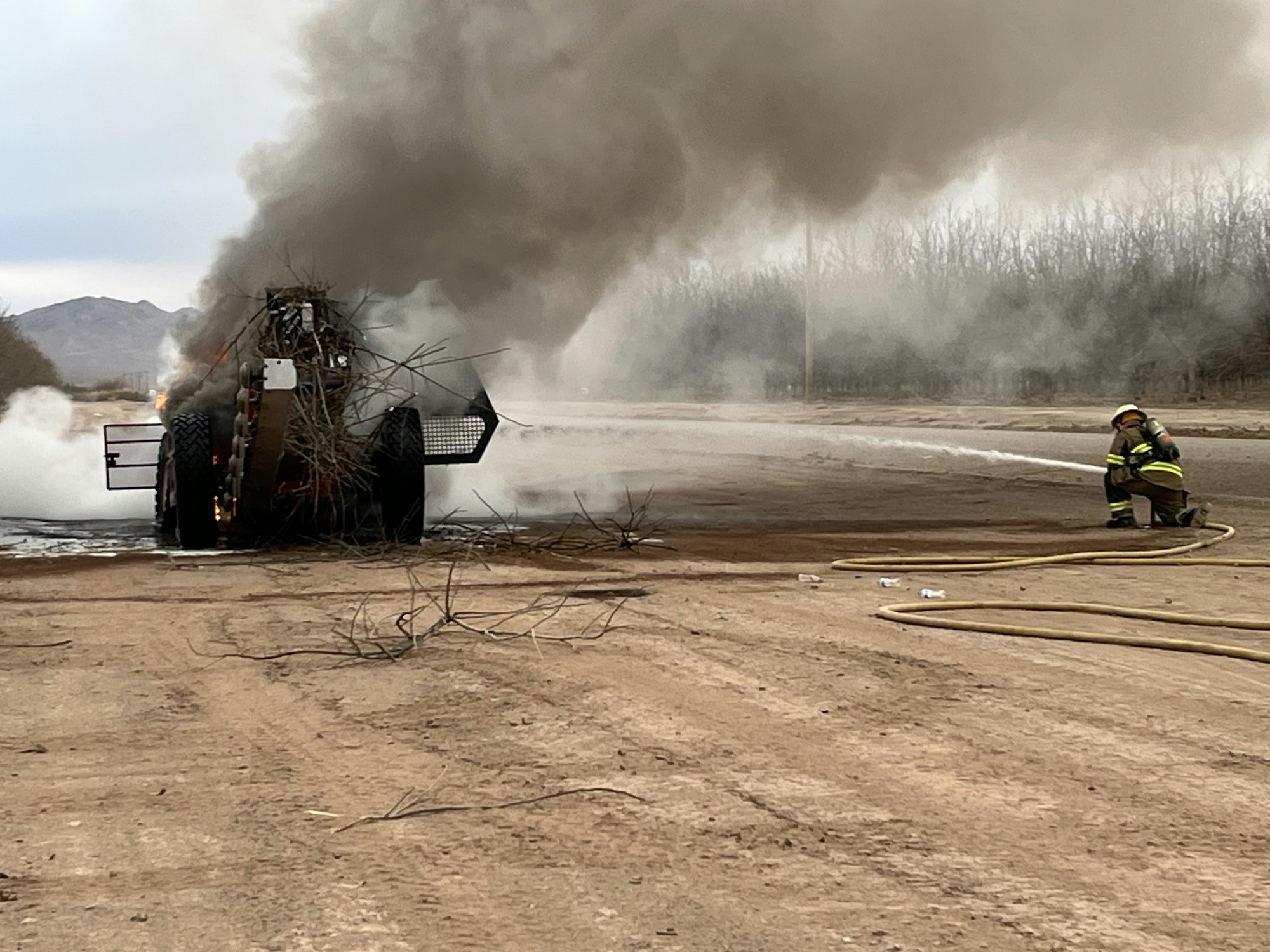 A firefighter is extinguishing a fire on a vehicle or equipment on a dirt road, with smoke and flames visible.