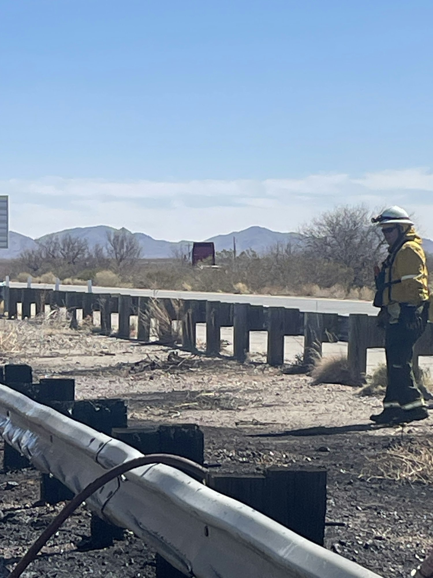 A person in protective gear stands near a roadside barrier under a clear sky.