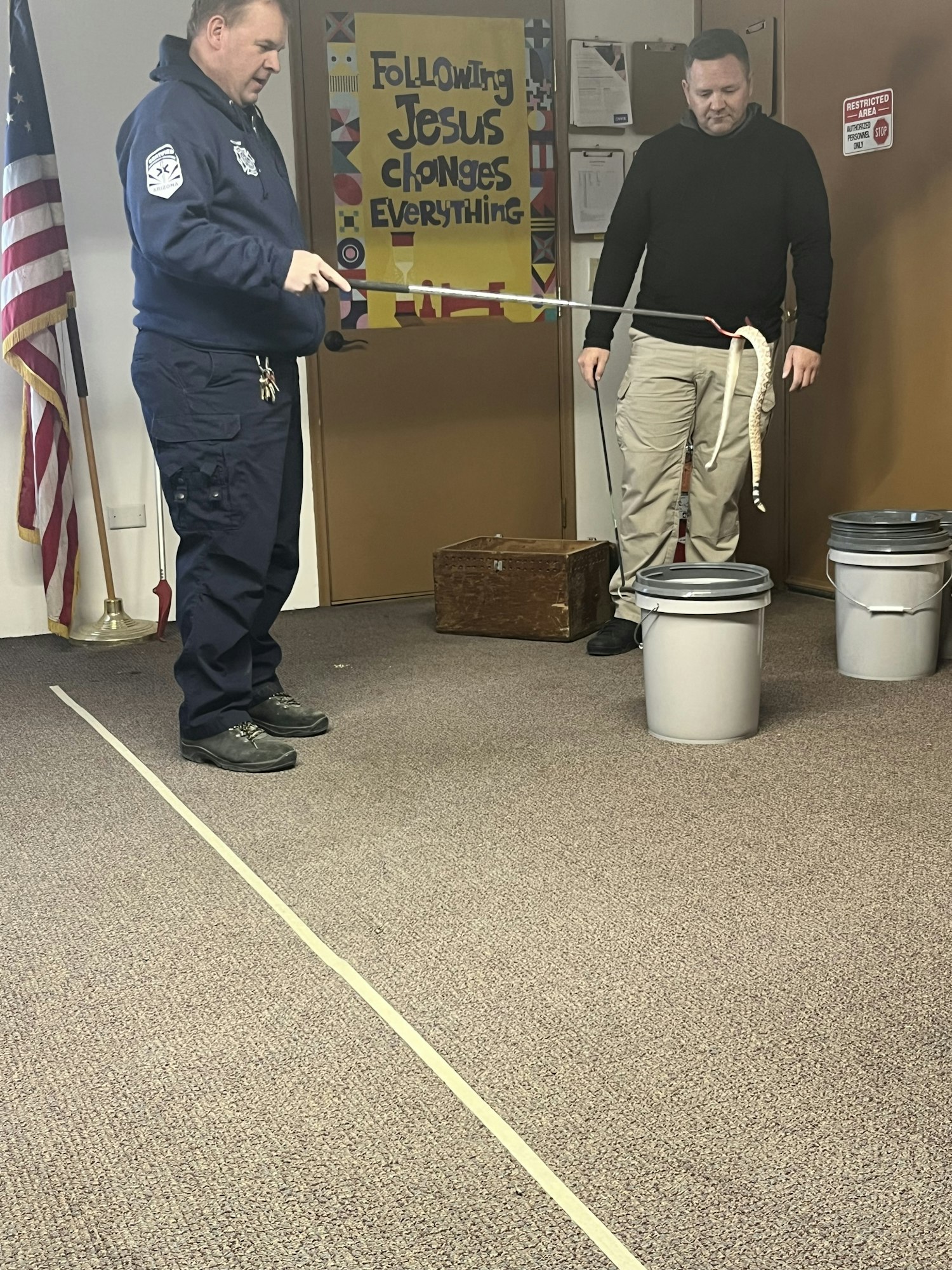 Two men handling a pale snake with a tool, near buckets, on carpeted floor. A sign in the background.