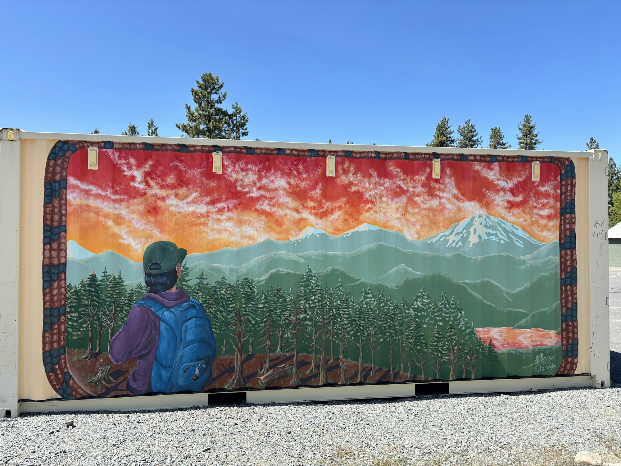 Painting on a container shows a person with a backpack facing a sunset over mountains and a forest.