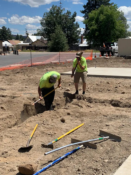 Two workers in bright shirts are digging in a dirt site, surrounded by tools and construction barriers, under a blue sky.