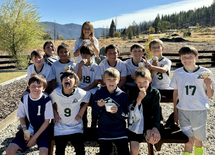 A group of cheerful kids in sports uniforms eating ice cream outdoors with mountains in the background.