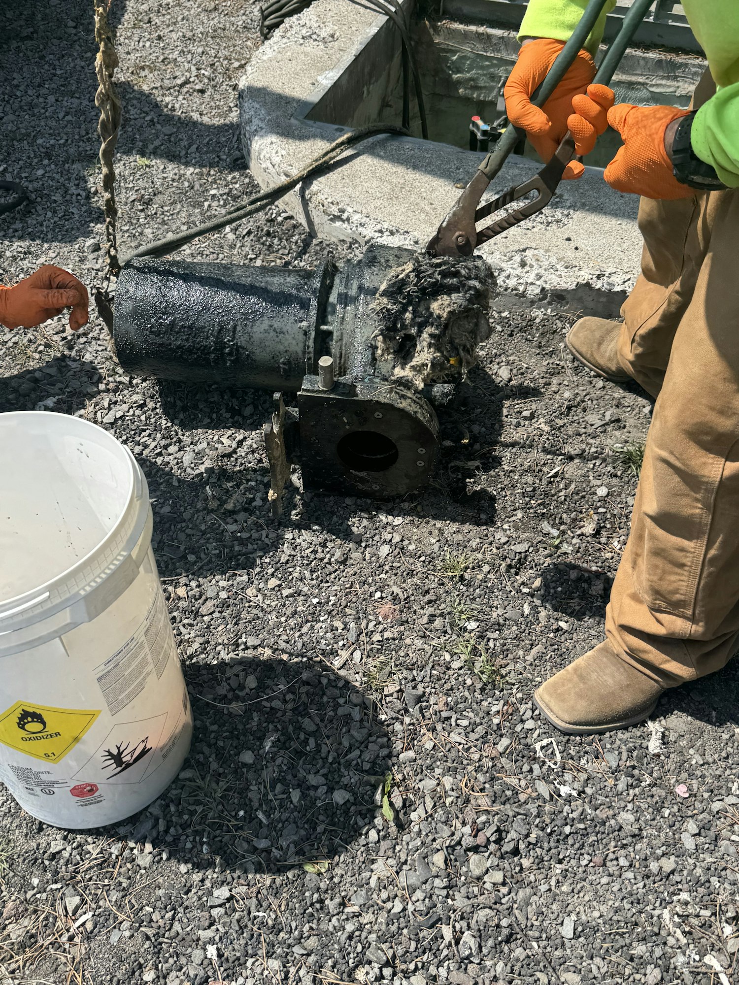 A person using pliers to remove debris from a pipe, with a hazardous waste bucket nearby on a gravel surface.