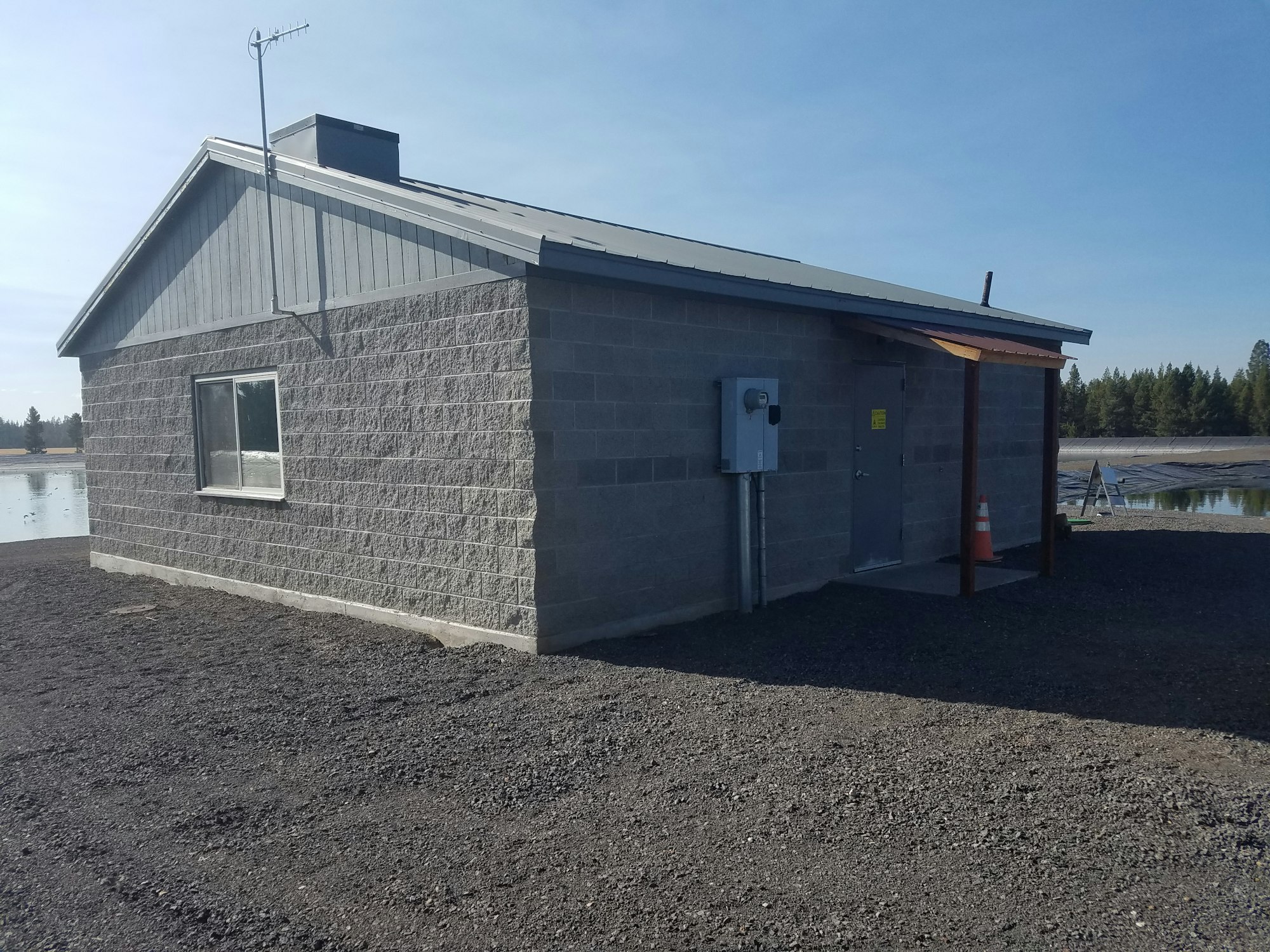 Small building with textured walls, metal roof, antenna, and an electrical box outside, near a body of water.