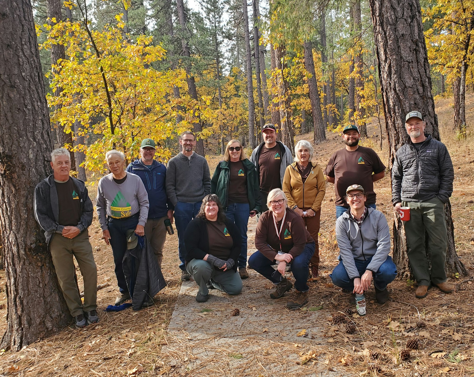 A group of people poses for a photo in a forest with autumn foliage, many wearing matching shirts.