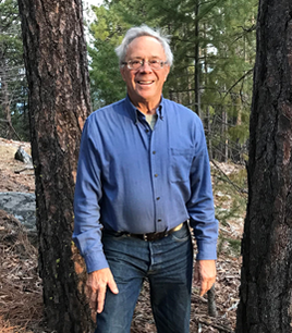 A man in a blue shirt and jeans stands between trees in a forested area, smiling.