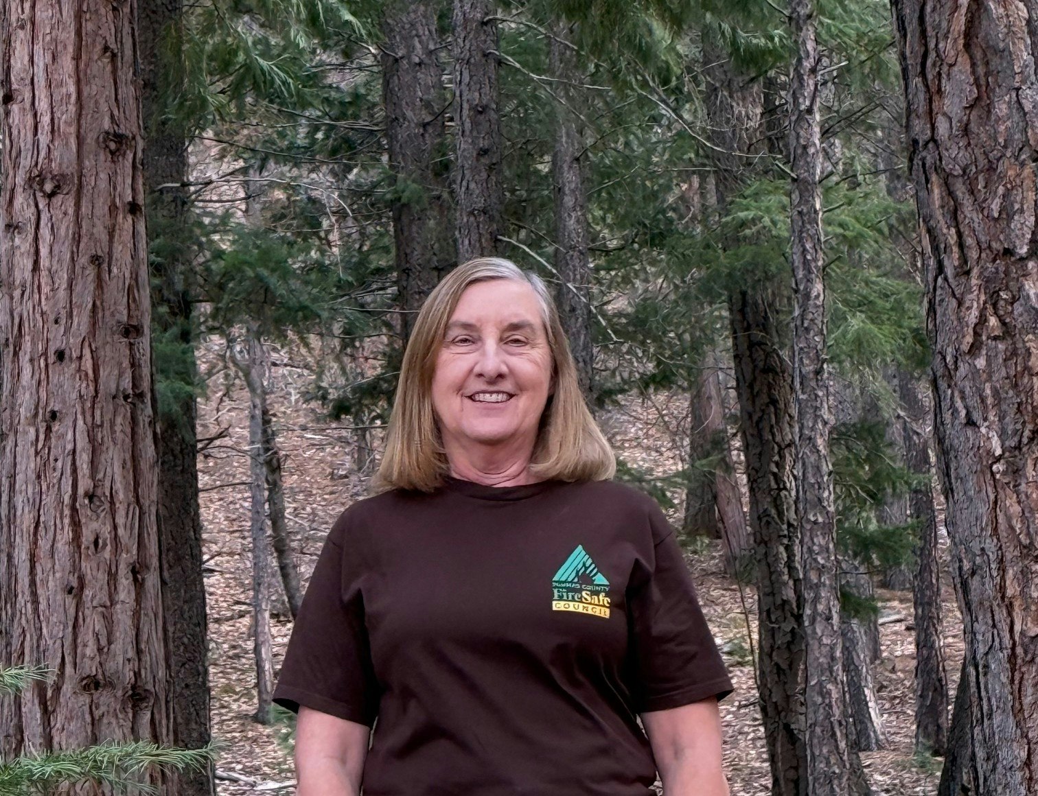 A woman smiles in a forest, wearing a brown shirt with a FireSafe Council logo, surrounded by trees.