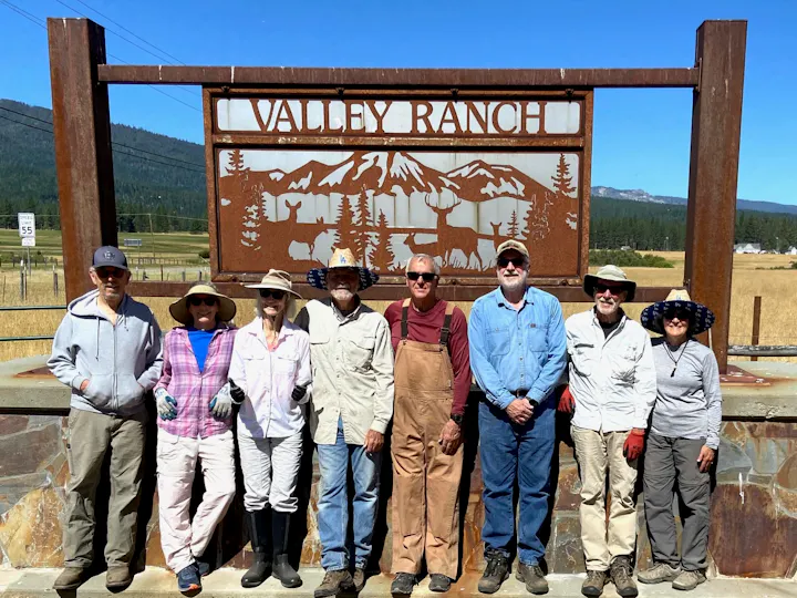 A group of eight people stands by a "Valley Ranch" sign in a scenic, outdoor setting. They're dressed for warm, sunny weather.