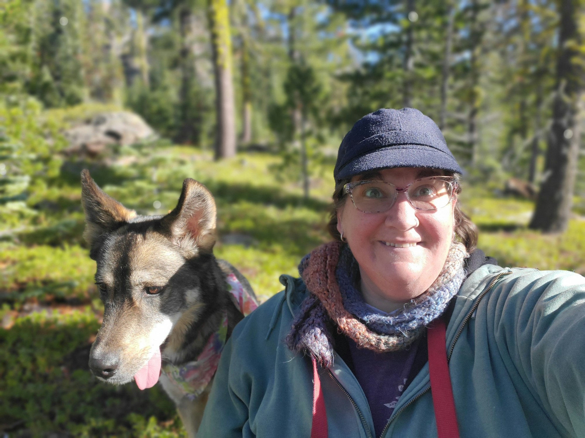 A person smiling with glasses, wearing a scarf and hat, posing outdoors with a dog in a natural setting.