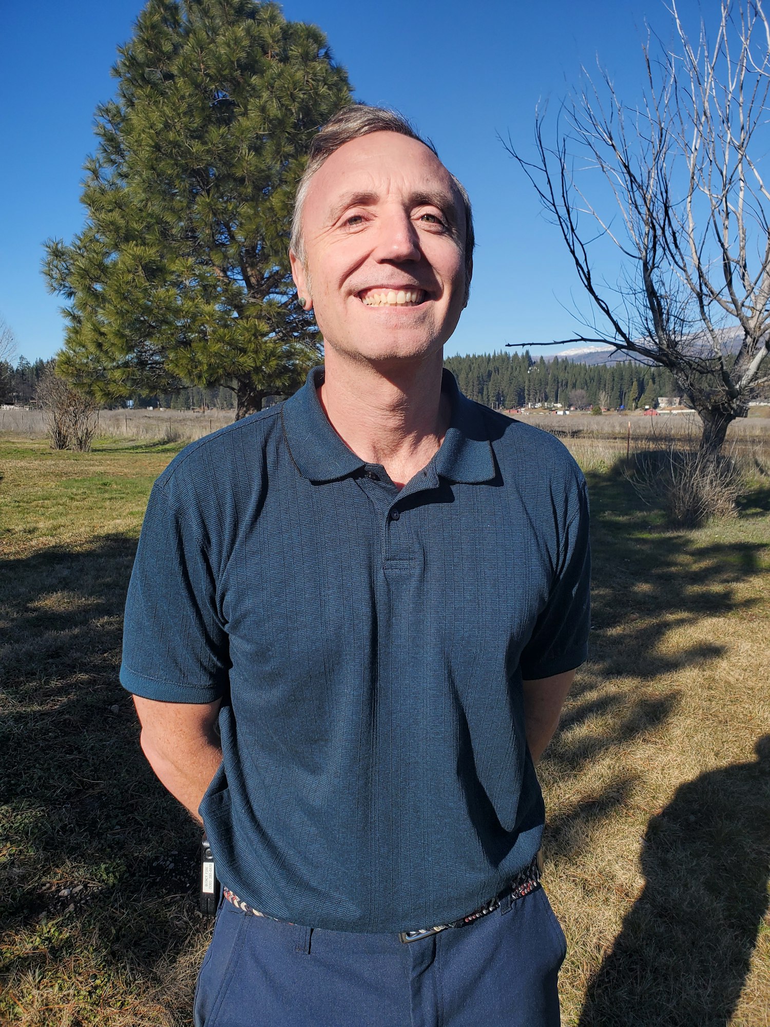 A smiling man in a dark polo shirt stands outdoors with trees and blue sky in the background.