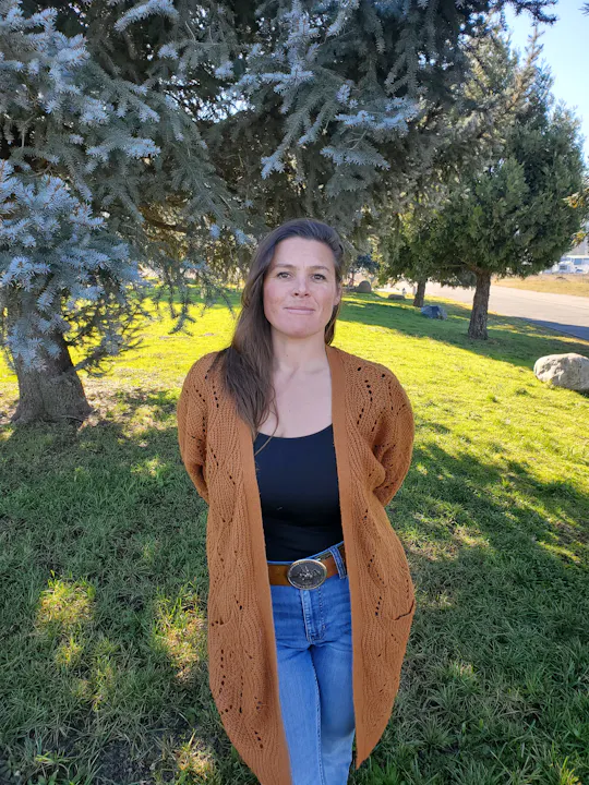 A woman stands outdoors in a cozy brown cardigan and jeans, surrounded by greenery and trees in a sunny setting.