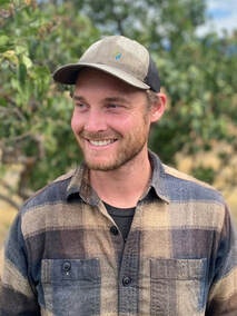 A man smiling, wearing a plaid shirt and cap, standing outdoors with trees in the background.