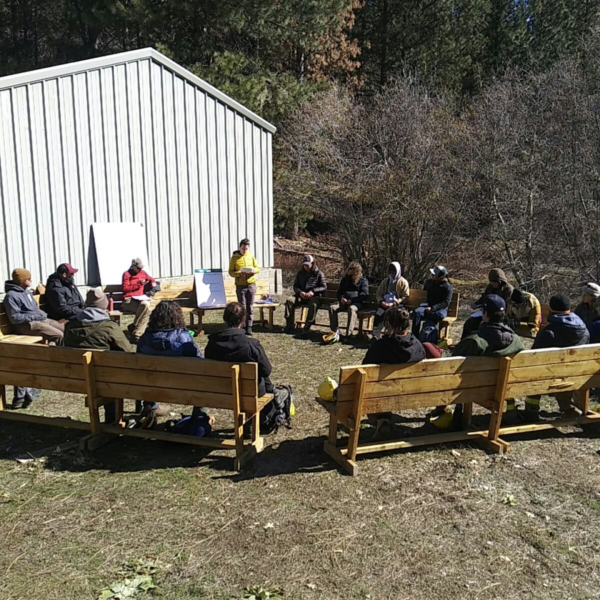 Outdoor meeting with people sitting on benches in a circle around a speaker with a flip chart near a building.