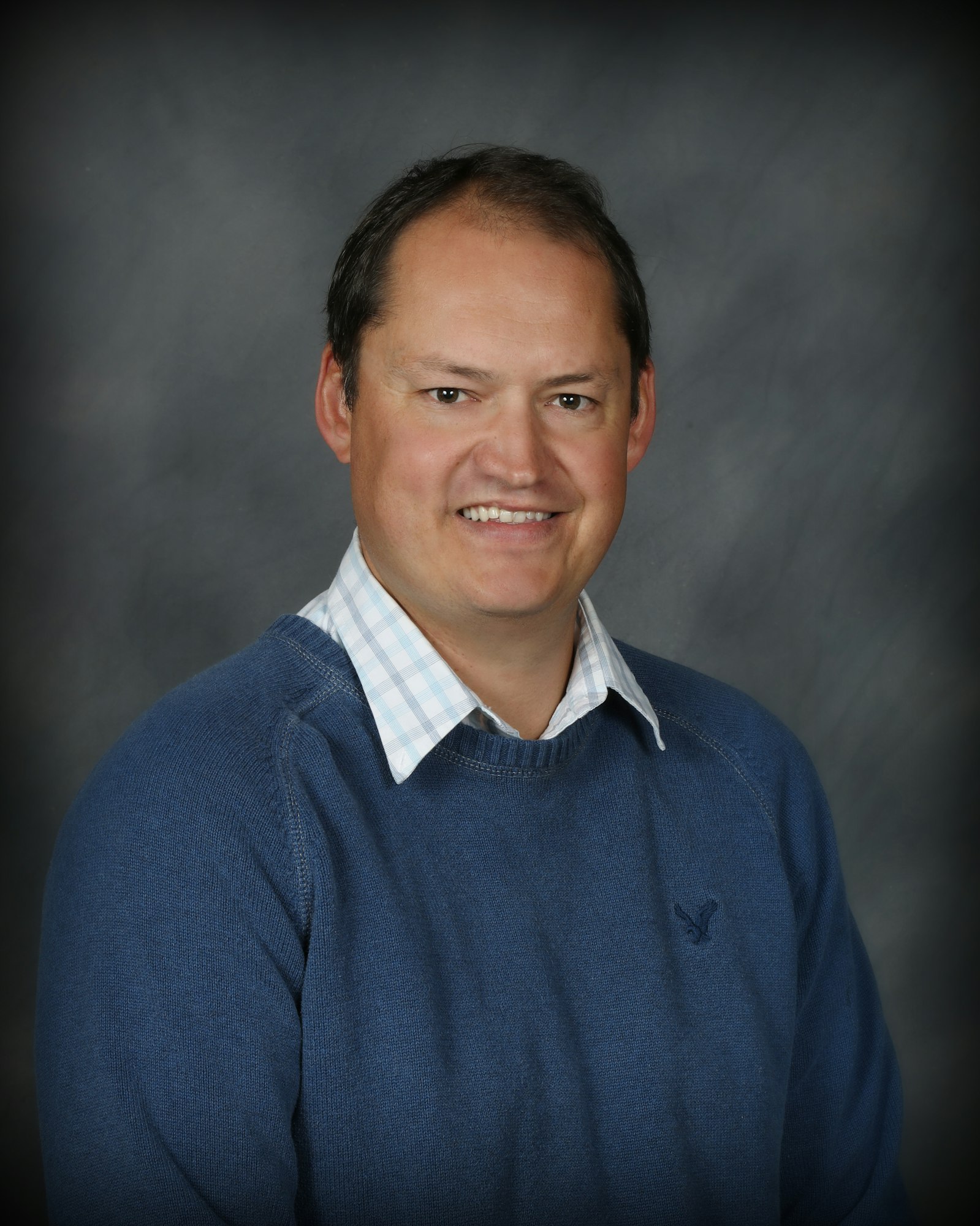 The image shows a smiling man wearing a blue sweater over a checkered shirt, against a gray background.