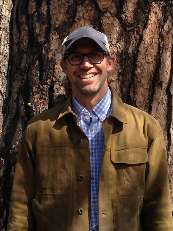 A person wearing glasses, a cap, and a brown jacket stands smiling in front of a tree.