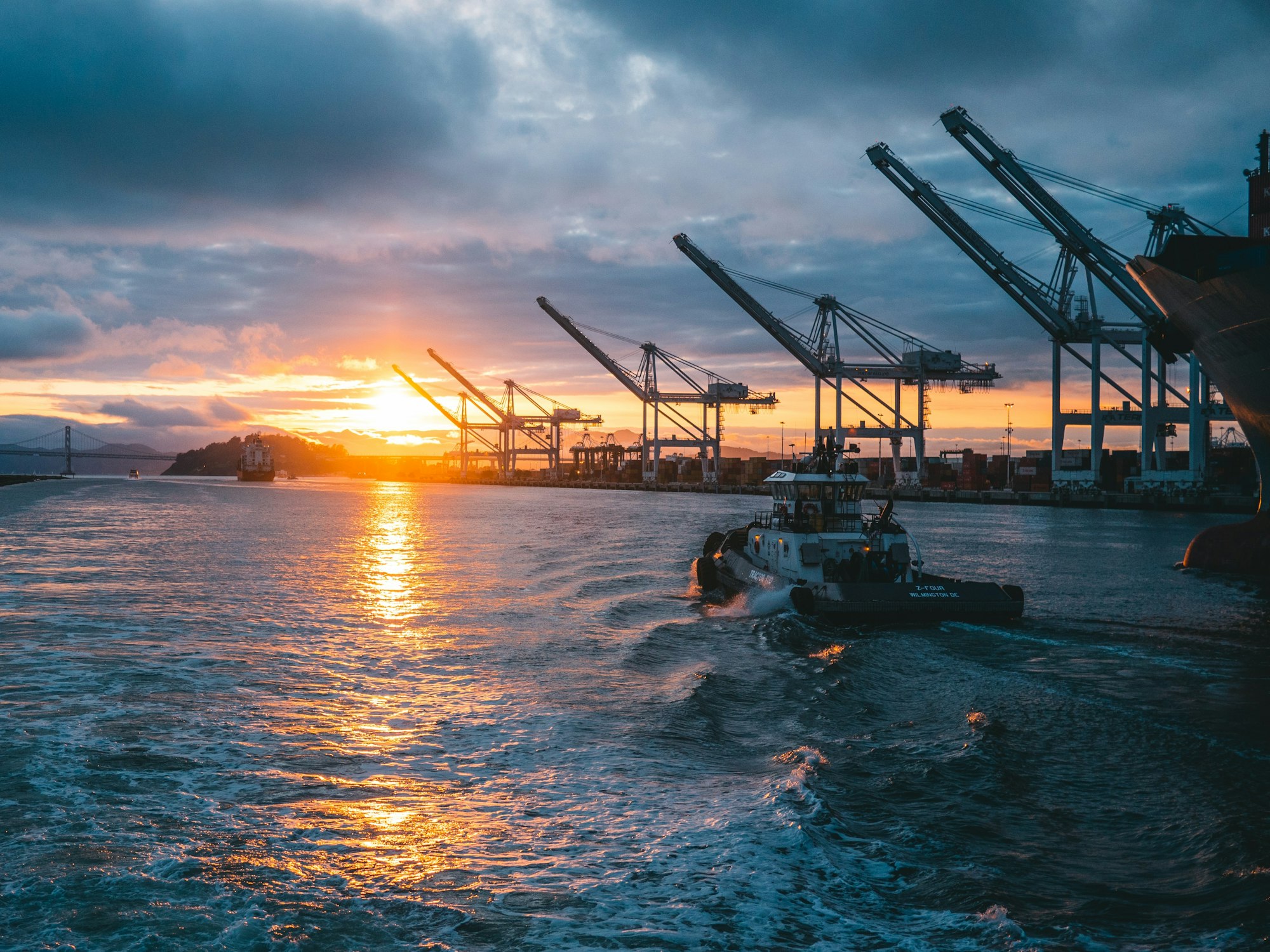 A tugboat navigates water near a port at sunset, with cranes and a distant bridge silhouetted against the colorful sky.