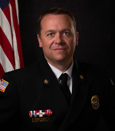 A person in a formal uniform with badges, an American flag in the background.