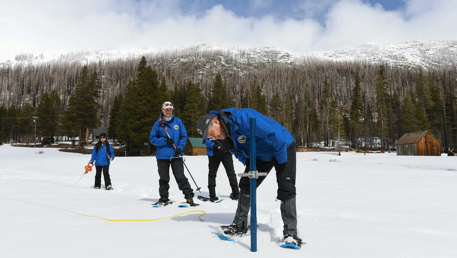 People in blue jackets measuring snow depth in a snowy landscape with trees and mountains in the background.