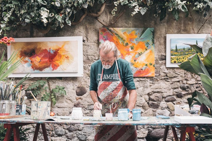 An artist works at a table with paint and brushes, surrounded by colorful paintings in a lush outdoor setting.