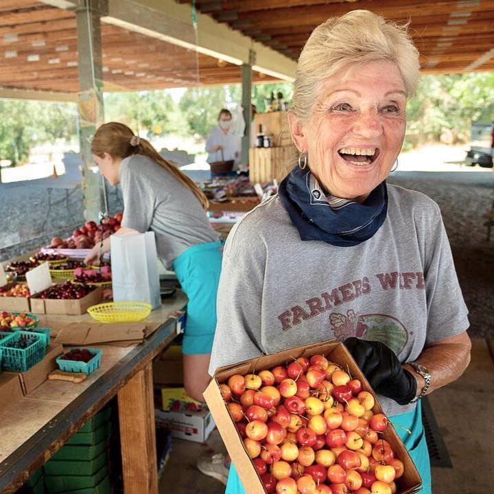 Smiling woman at a market stall holding a box of cherries; others sorting fruit in the background.