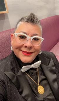 Smiling person with short hair, white glasses, in a suit with a bow tie and necklace, sitting on a purple couch.
