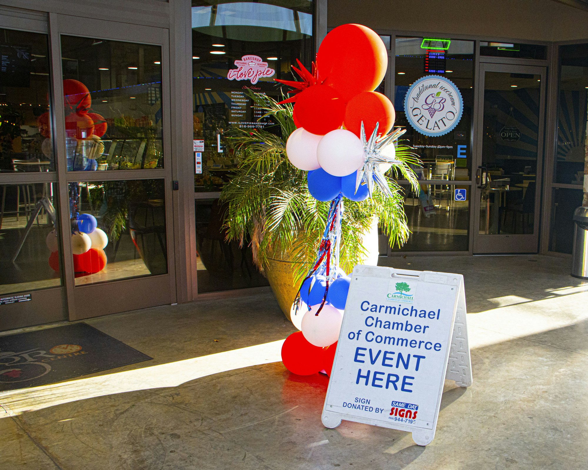 Sign for "Carmichael Chamber of Commerce Event Here" with red, white, and blue balloons outside a gelato shop.