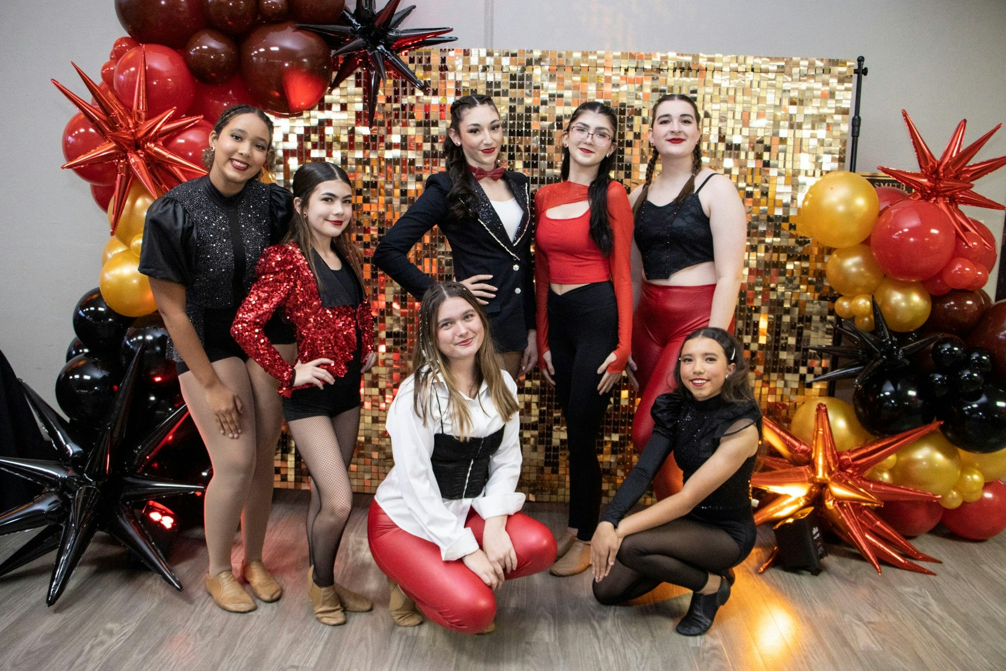 Seven people posing in dance costumes with a backdrop of gold sequins and red, gold, and black balloon decorations.