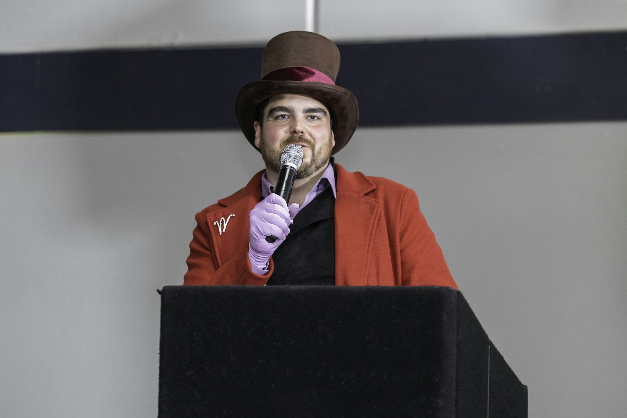 A person in a red coat and top hat, wearing a purple glove, speaks into a microphone at a podium.