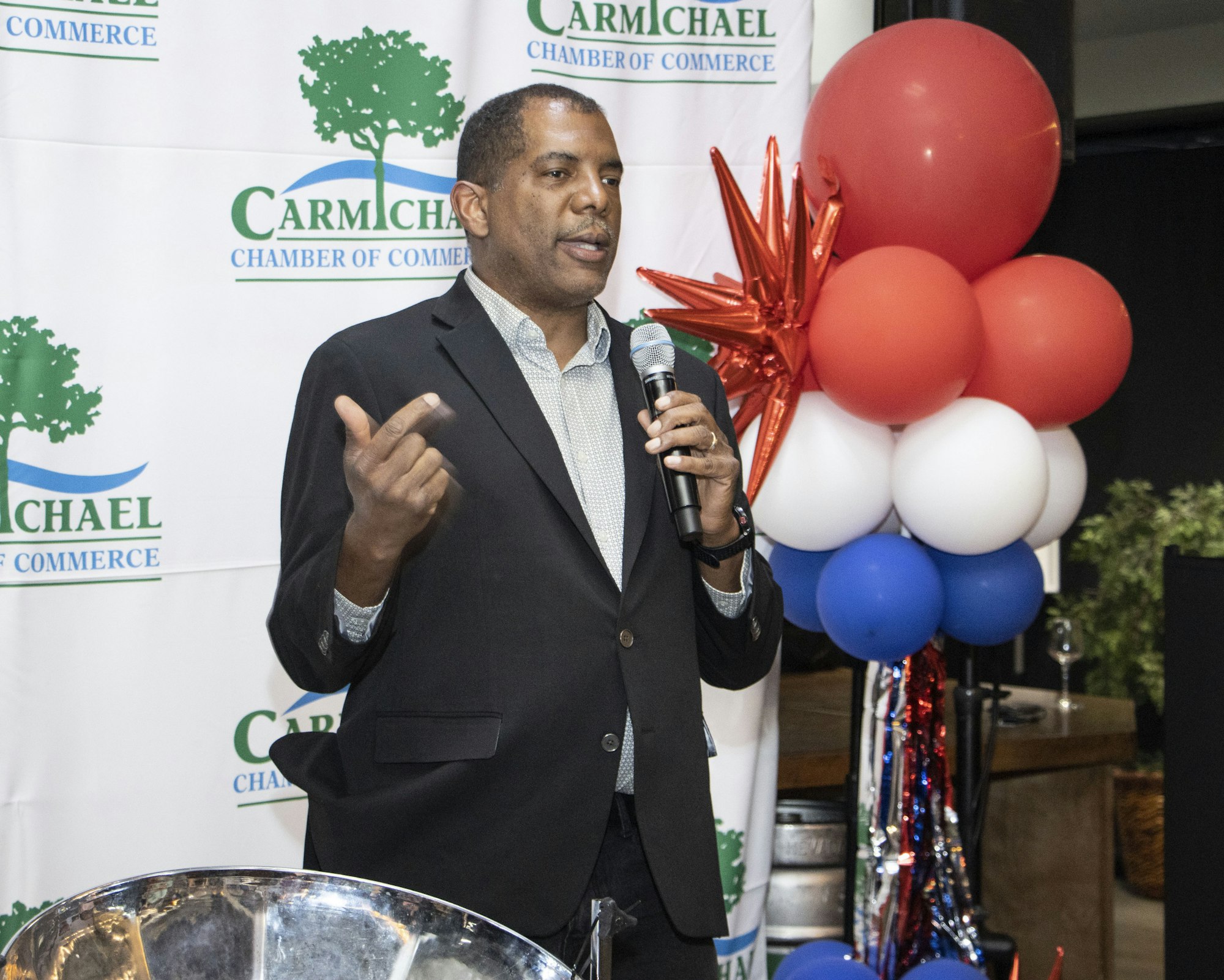 Person speaking at an event with a microphone, standing by a Carmichael Chamber of Commerce backdrop and red, white, and blue balloons.
