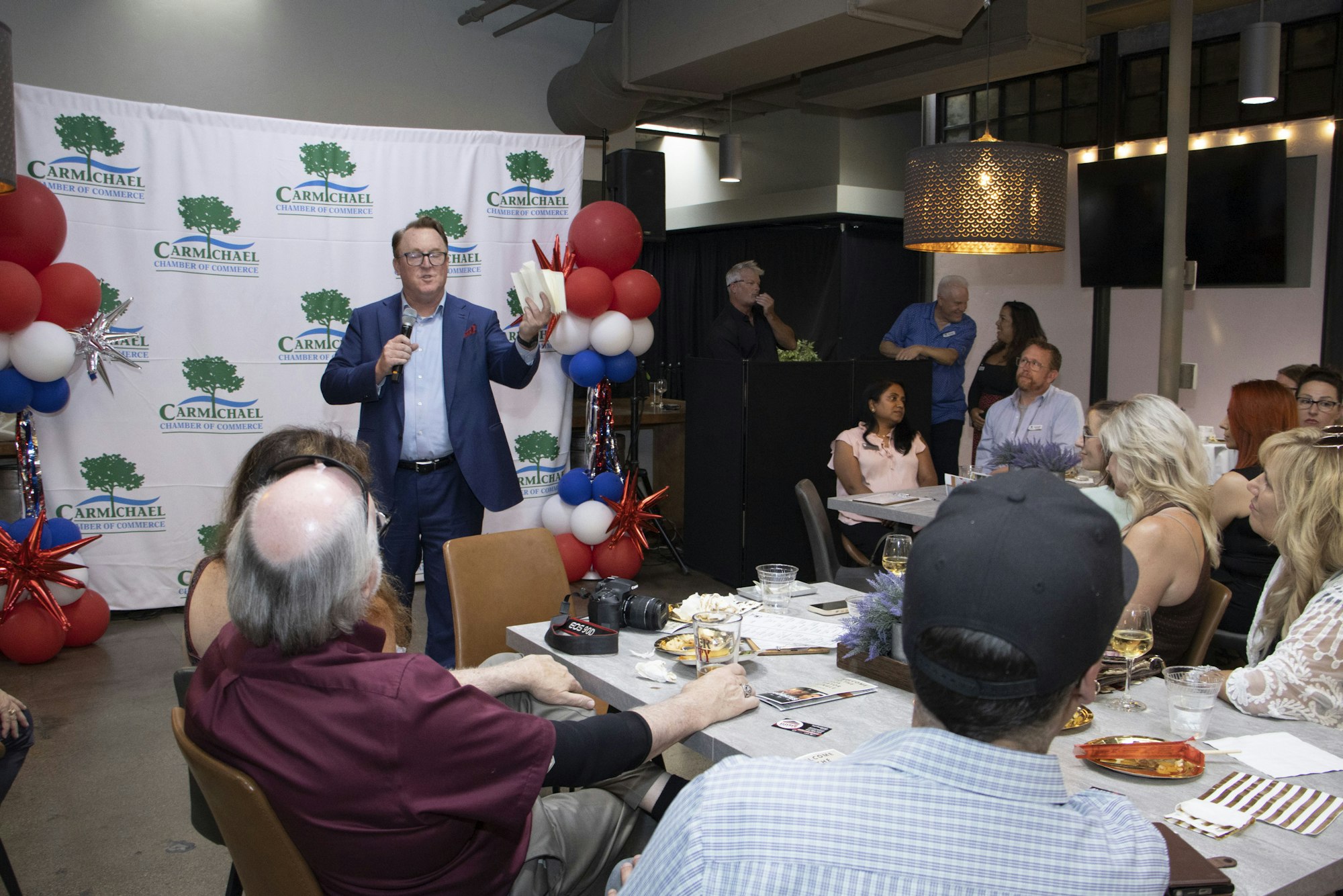 A man speaks to a seated audience at a Carmichael Chamber of Commerce event with balloons and a backdrop.