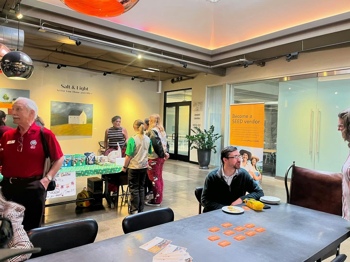 People interacting at a community event indoors, with tables, snacks, and informational displays.